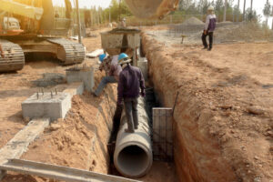 Workers install large concrete pipes in a trench with excavator and footings nearby.
