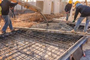 Workers are pouring concrete onto a large steel rebar grid, forming a continuous mat slab footing.