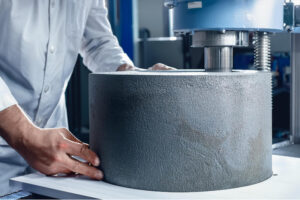 Person in lab coat places large gray cylinder under a compression testing machine.